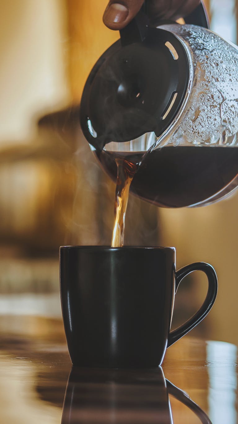 Steaming hot coffee being poured from a glass pot into a black mug, perfect morning drink.